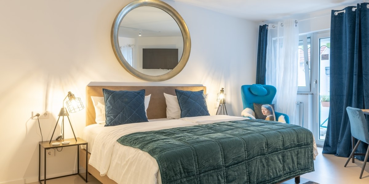 Modernes Doppelzimmer mit großem Bett, blauen Kissen und Decke, rundem Spiegel, blauen Vorhängen und Sessel., © Hotel Princess