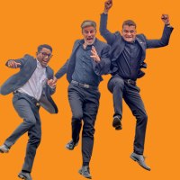 Drei Männer in Anzügen springen fröhlich in die Luft vor einem orangefarbenen Hintergrund. Sie wirken energiegeladen und begeistert., © Stadt Murrhardt