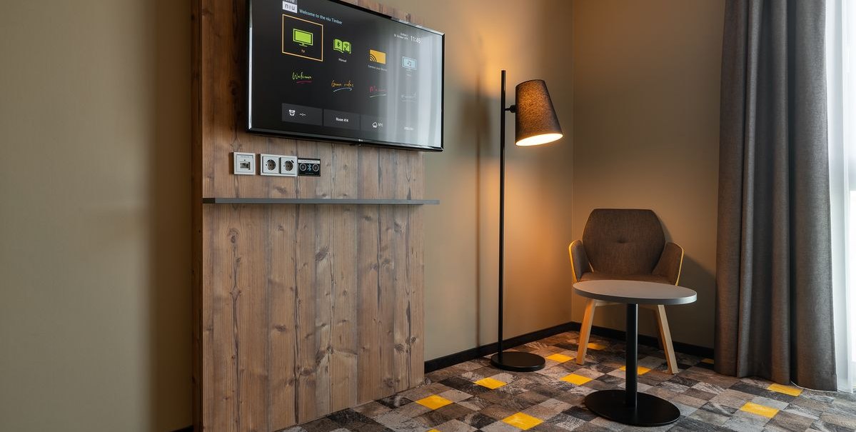 Modernes Hotelzimmer mit Holzverkleidung, Flachbildfernseher, Stehlampe, Sessel und kleinem Tisch auf gemustertem Teppichboden., © Novum Hospitality Modernes Hotelzimmer mit Holzverkleidung, Flachbildfernseher, Stehlampe, Sessel und kleinem Tisch auf gemustertem Teppichboden., © Novum Hospitality