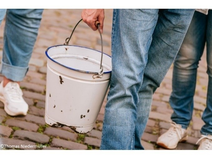 Person in Jeans tr&auml;gt einen wei&szlig;en Eimer auf Kopfsteinpflaster. Zwei weitere Personen sind teilweise sichtbar., &copy; Stuttgart Marketing GmbH