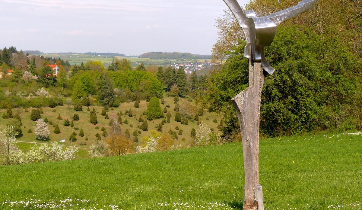Eine moderne Skulptur steht auf einer gr&uuml;nen Wiese mit Blick auf eine h&uuml;gelige Landschaft und bl&uuml;hende B&auml;ume., &copy; Natur.Nah. Sch&ouml;nbuch & Heckeng&auml;u