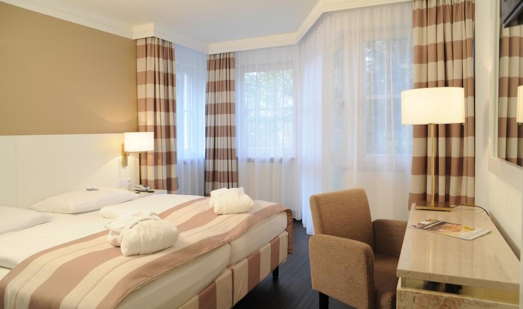 Elegantes Hotelzimmer mit Doppelbett, gestreiften Vorhängen und einem Schreibtisch. Helle, einladende Atmosphäre mit moderner Einrichtung., © relexa Waldhotel Schatten Elegantes Hotelzimmer mit Doppelbett, gestreiften Vorhängen und einem Schreibtisch. Helle, einladende Atmosphäre mit moderner Einrichtung., © relexa Waldhotel Schatten