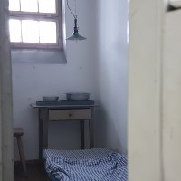 Blick in eine historische Gefängniszelle mit einfachem Bett, kleinem Tisch und Schüsseln. Ein Fenster mit Gitterstäben lässt Licht herein., © Strafvollzugsmuseum Ludwigsburg