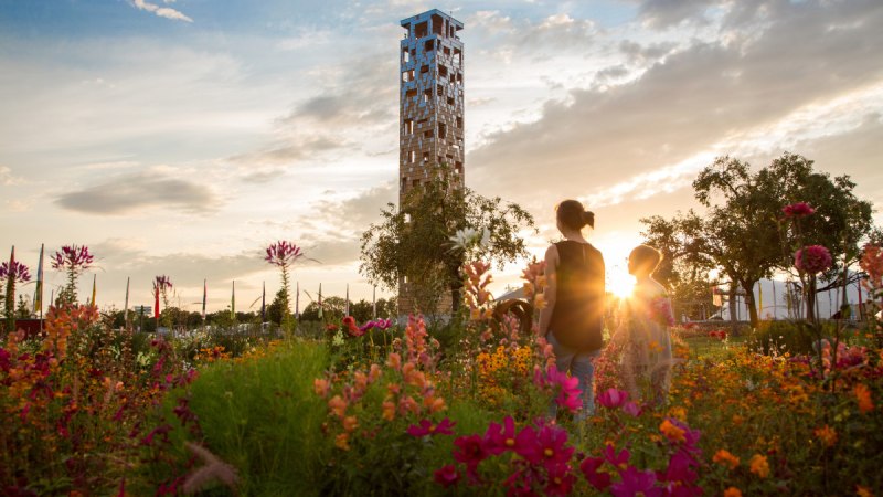 Ein Turm im Himmelsgarten, umgeben von bunten Blumen. Zwei Personen stehen im Vordergrund, w&auml;hrend die Sonne im Hintergrund untergeht., &copy; Foto Thomas Zehnder