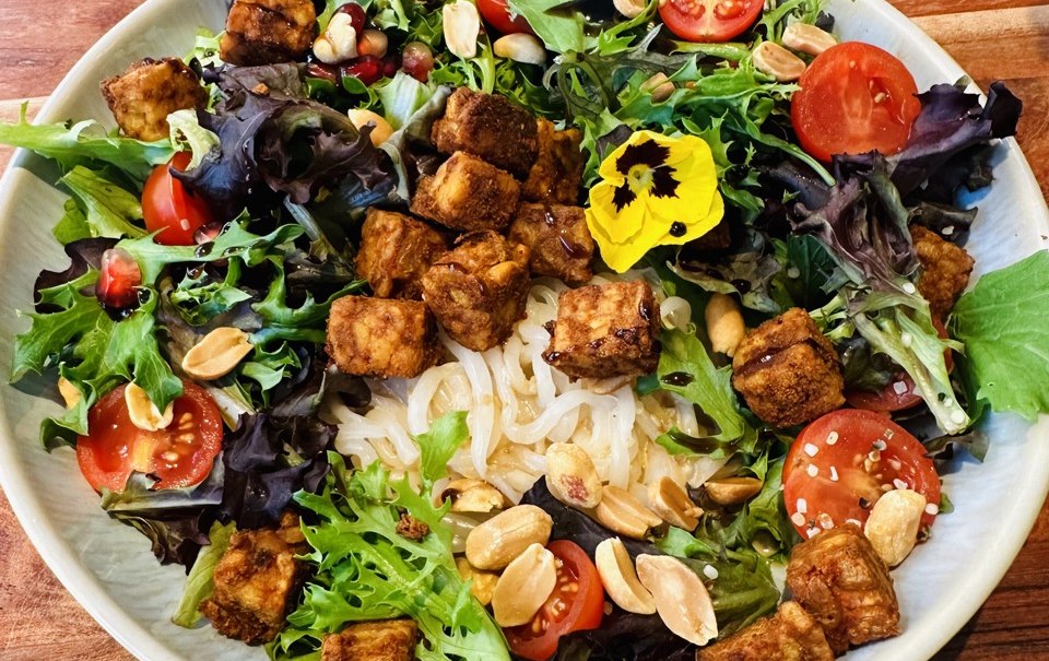 Ein bunter Salat mit Tofu, Kirschtomaten, Erdn&uuml;ssen und essbaren Bl&uuml;ten auf einem Holztisch. Besteck liegt auf einer Serviette daneben., &copy; cStadtN&uuml;rtingen