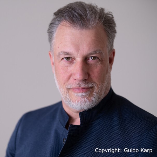 Ein Mann mit grauem Haar und Bart trägt eine dunkle Jacke und blickt direkt in die Kamera vor einem neutralen Hintergrund., © MW Promotion GmbH