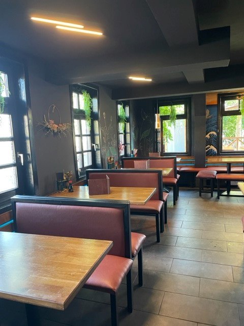 Gemütliches Restaurant mit Holztischen und roten Sitzbänken. Dunkle Wände und Fenster mit Pflanzen sorgen für eine einladende Atmosphäre., © cAvada Gemütliches Restaurant mit Holztischen und roten Sitzbänken. Dunkle Wände und Fenster mit Pflanzen sorgen für eine einladende Atmosphäre., © cAvada