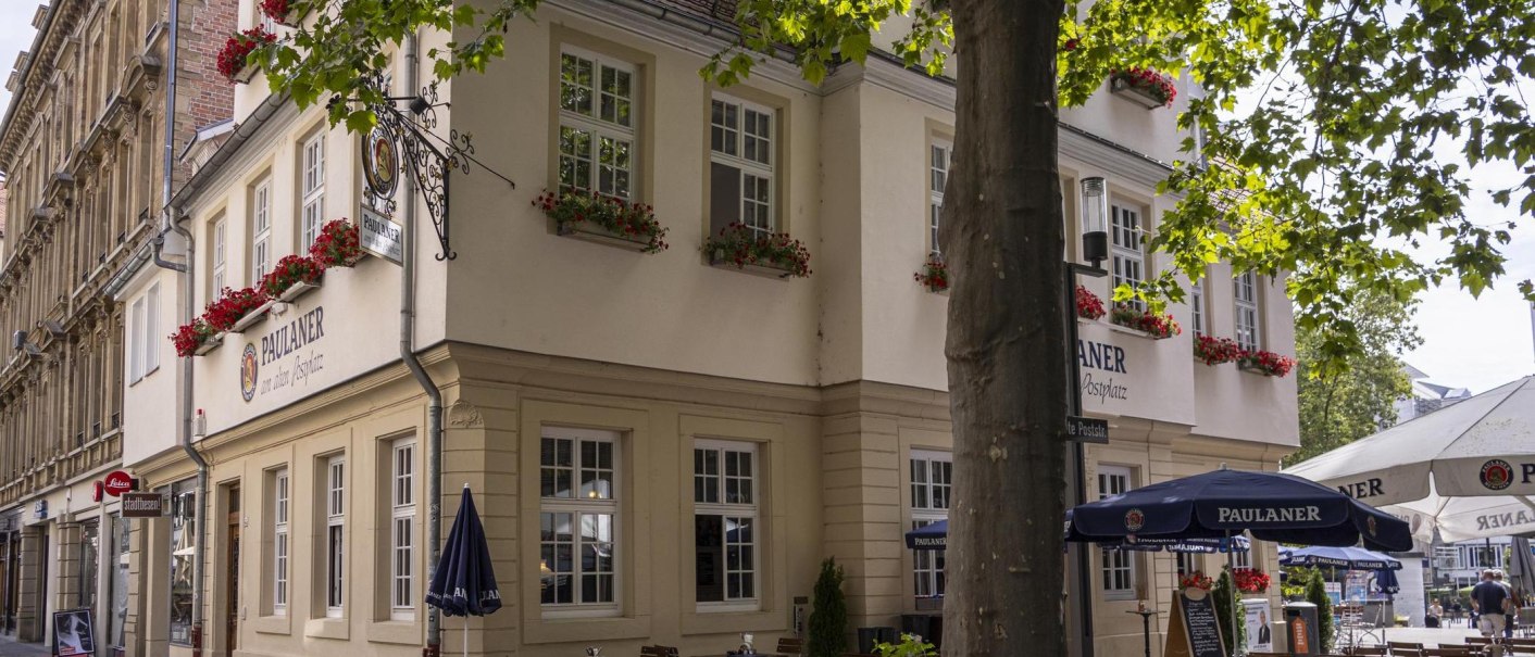 Eckgebäude mit Paulaner-Restaurant, dekoriert mit roten Blumen. Vor dem Gebäude stehen Sonnenschirme und Tische. Bäume spenden Schatten., © SMG, Sarah Schmid Eckgebäude mit Paulaner-Restaurant, dekoriert mit roten Blumen. Vor dem Gebäude stehen Sonnenschirme und Tische. Bäume spenden Schatten., © SMG, Sarah Schmid