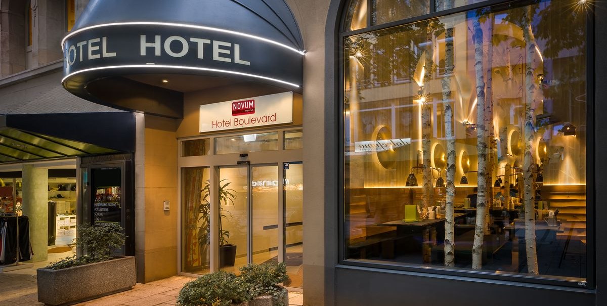 Außenansicht eines Hotels mit beleuchtetem Eingang, großem Fenster und Pflanzen. Schild mit der Aufschrift 'Hotel Boulevard' über der Tür., © Novum Hospitality