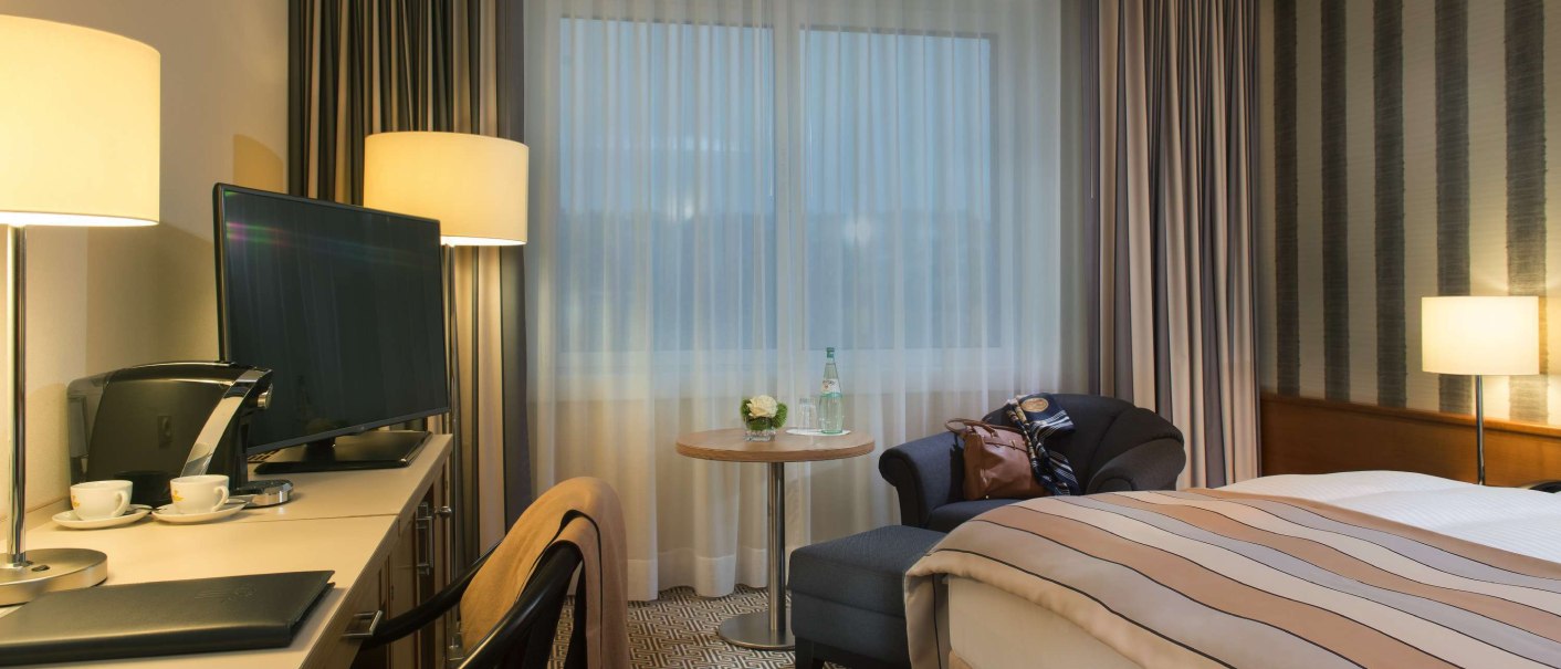 Modernes Hotelzimmer mit Doppelbett, Schreibtisch, Fernseher und gemütlicher Beleuchtung. Vorhänge und Teppich ergänzen das stilvolle Ambiente., © Maritim Hotelgesellschaft mbH