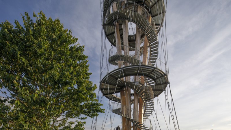 Ein spiralförmiger Aussichtsturm aus Holz und Metall erhebt sich neben einem grünen Baum unter einem blauen Himmel., © Stoppel
