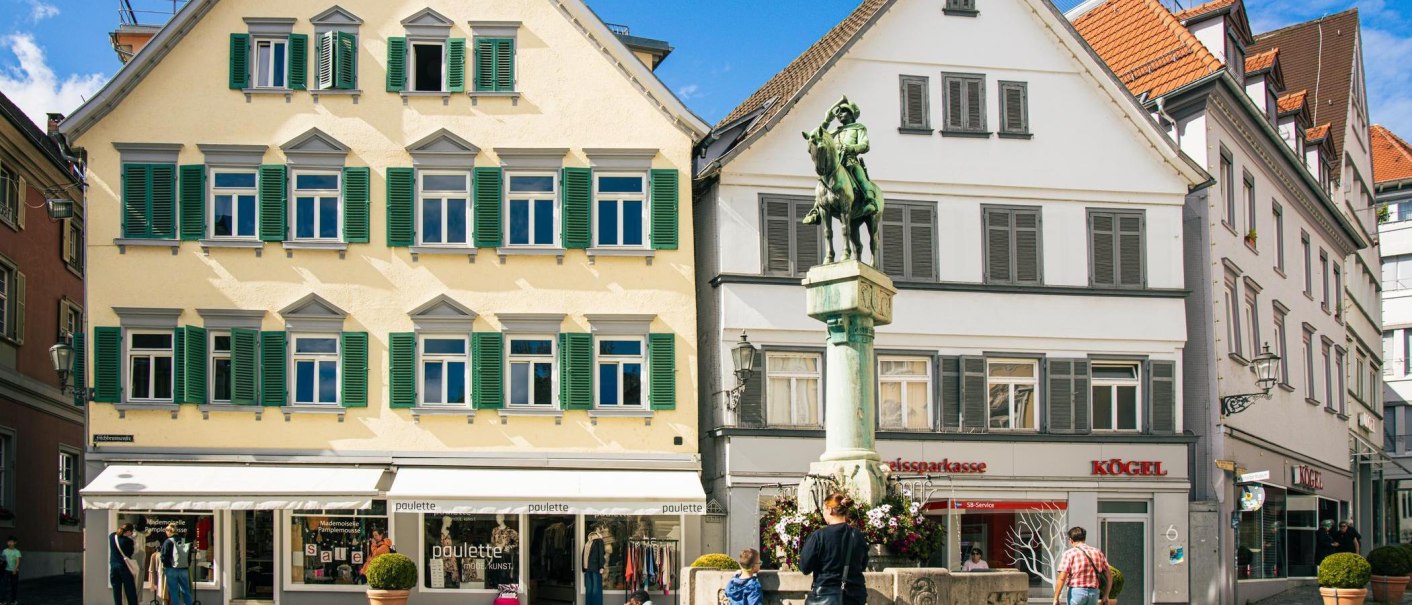 Der Postmichelbrunnen auf dem Marktplatz in Esslingen, flankiert von historischen Gebäuden mit Geschäften im Erdgeschoss. Menschen spazieren vorbei., © Stuttgart-Marketing GmbH, Sarah Schmid Der Postmichelbrunnen auf dem Marktplatz in Esslingen, flankiert von historischen Gebäuden mit Geschäften im Erdgeschoss. Menschen spazieren vorbei., © Stuttgart-Marketing GmbH, Sarah Schmid