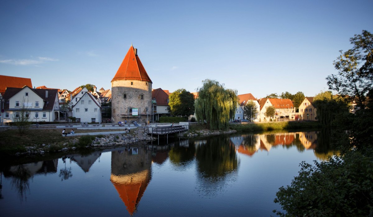 Ein mittelalterlicher Turm mit rotem Dach und umliegende Häuser spiegeln sich in einem ruhigen Fluss. Die Szene ist von Bäumen umgeben., © Stoppel