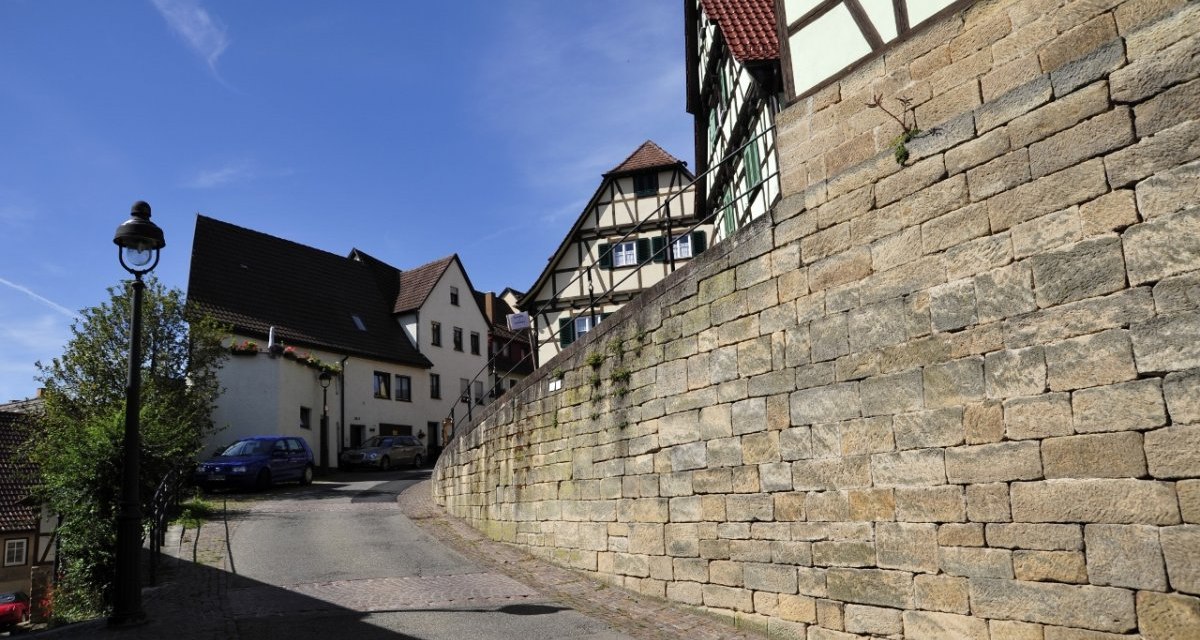Gepflasterte Stra&szlig;e mit Fachwerkh&auml;usern und einer Steinmauer, blauer Himmel, Laterne und Autos im Hintergrund., &copy; Natur.Nah. Sch&ouml;nbuch & Heckeng&auml;u