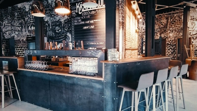 Moderne Bar mit industriellem Design, hohen Stühlen und Wandkunst. Gläser stehen auf der Theke, im Hintergrund eine Tafel mit Getränken., © Kraftpaule Moderne Bar mit industriellem Design, hohen Stühlen und Wandkunst. Gläser stehen auf der Theke, im Hintergrund eine Tafel mit Getränken., © Kraftpaule