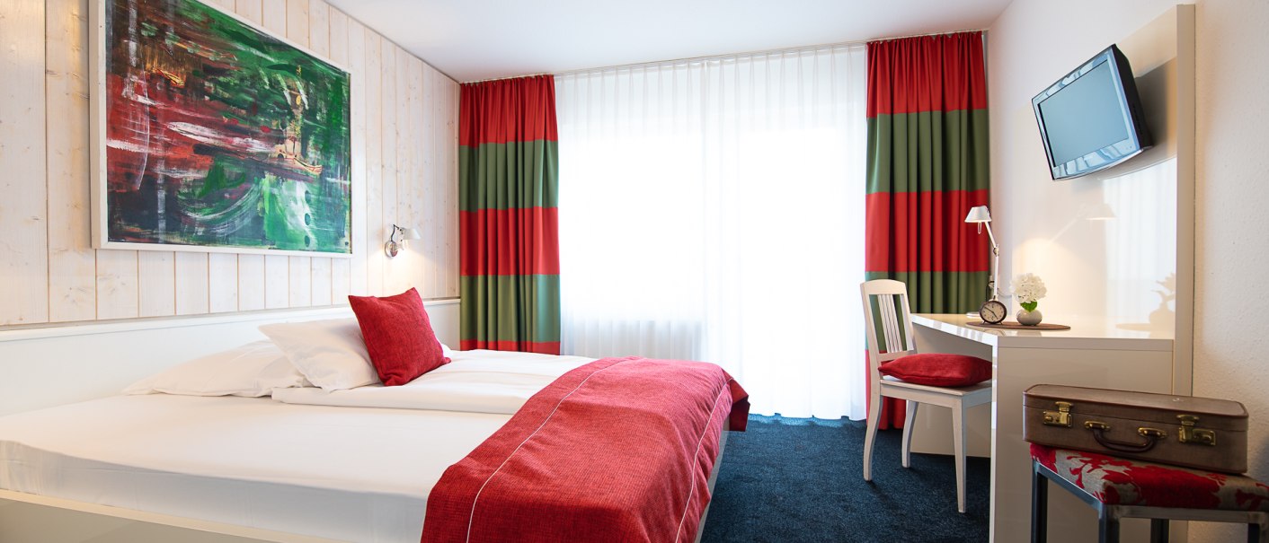 Gemütliches Zimmer mit Doppelbett, roter Bettwäsche, Schreibtisch, Stuhl und Gemälde. Vorhänge in Rot und Grün, TV an der Wand, Koffer auf Hocker., © Hotel Weinstadt