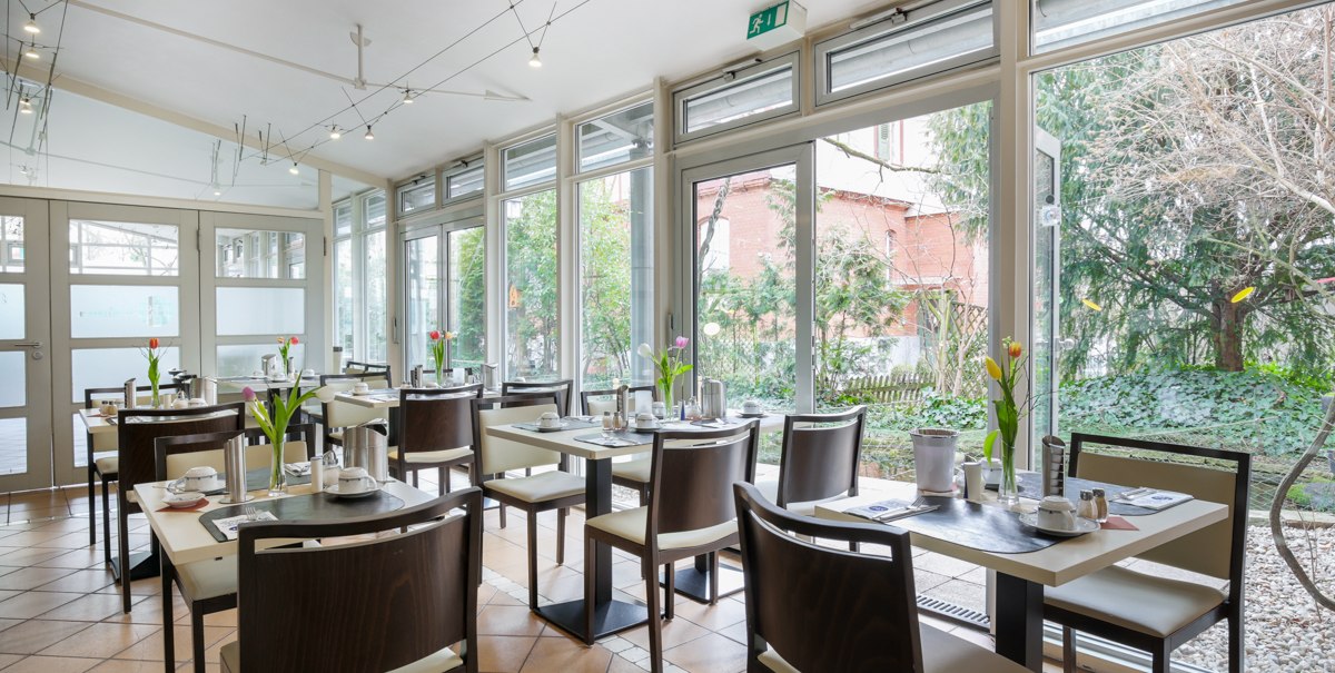 Helles Restaurant mit großen Fenstern, gedeckten Tischen und Blick auf einen Garten. Moderne Einrichtung mit schlichten Stühlen und Tischen., © TOMAS Helles Restaurant mit großen Fenstern, gedeckten Tischen und Blick auf einen Garten. Moderne Einrichtung mit schlichten Stühlen und Tischen., © TOMAS