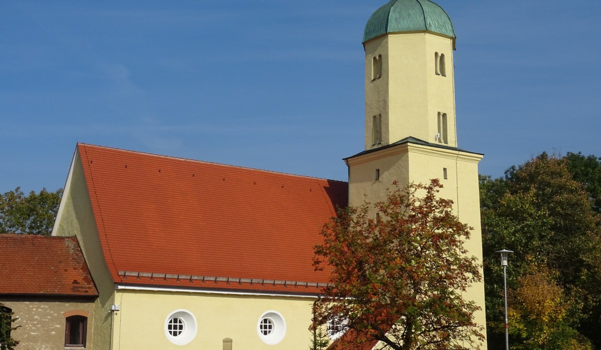 Die Dorfkirche Lauterburg mit gelbem Turm und rotem Dach steht vor einem klaren blauen Himmel, umgeben von Herbstbäumen., © Foto: Cornelia Steinbach Die Dorfkirche Lauterburg mit gelbem Turm und rotem Dach steht vor einem klaren blauen Himmel, umgeben von Herbstbäumen., © Foto: Cornelia Steinbach