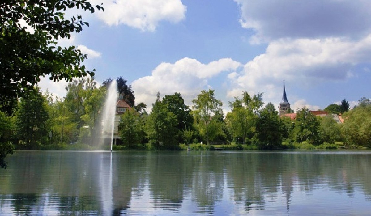 Ein idyllischer See mit einem Springbrunnen, umgeben von Bäumen. Im Hintergrund ist ein Kirchturm zu sehen, der in den blauen Himmel ragt., © Sindelfingen - Stuttgart-Marketing GmbH Ein idyllischer See mit einem Springbrunnen, umgeben von Bäumen. Im Hintergrund ist ein Kirchturm zu sehen, der in den blauen Himmel ragt., © Sindelfingen - Stuttgart-Marketing GmbH