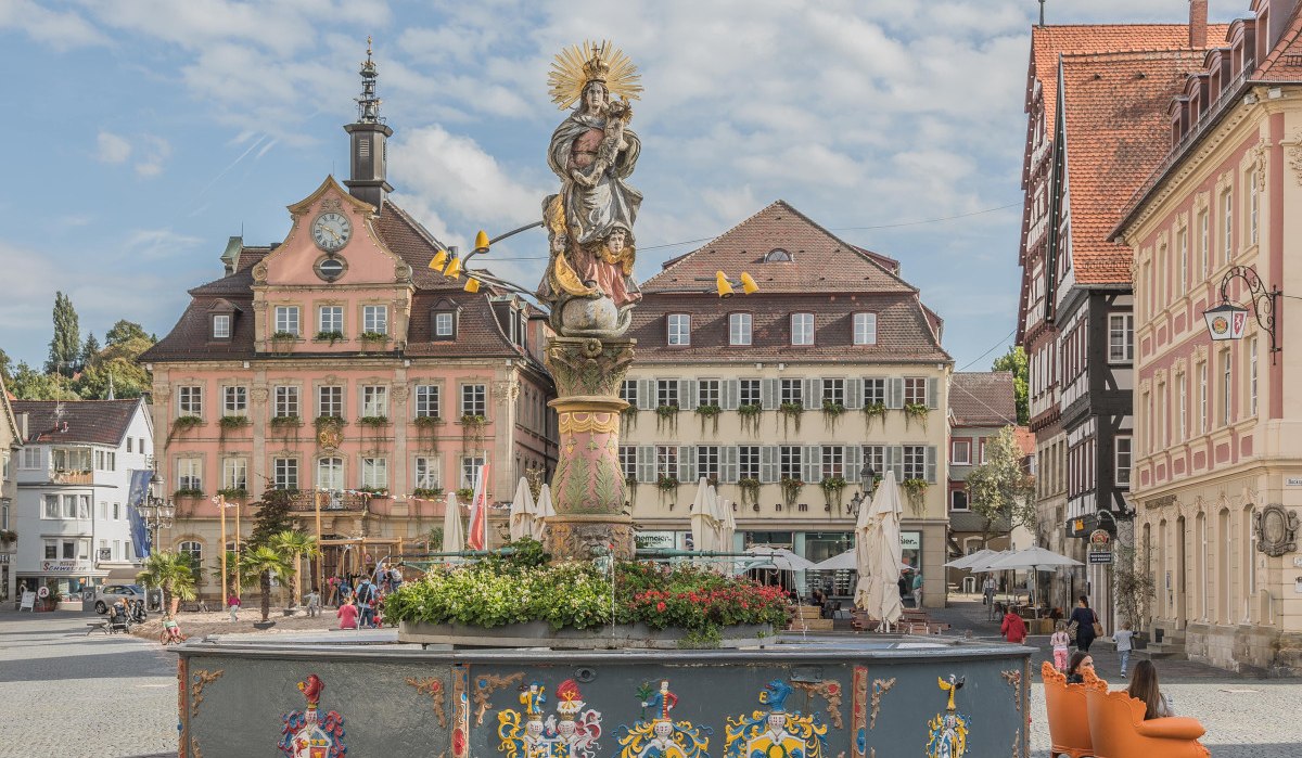 Historischer Marktplatz mit einem verzierten Brunnen und einem alten Rathaus im Hintergrund. Menschen flanieren und genie&szlig;en die Atmosph&auml;re.