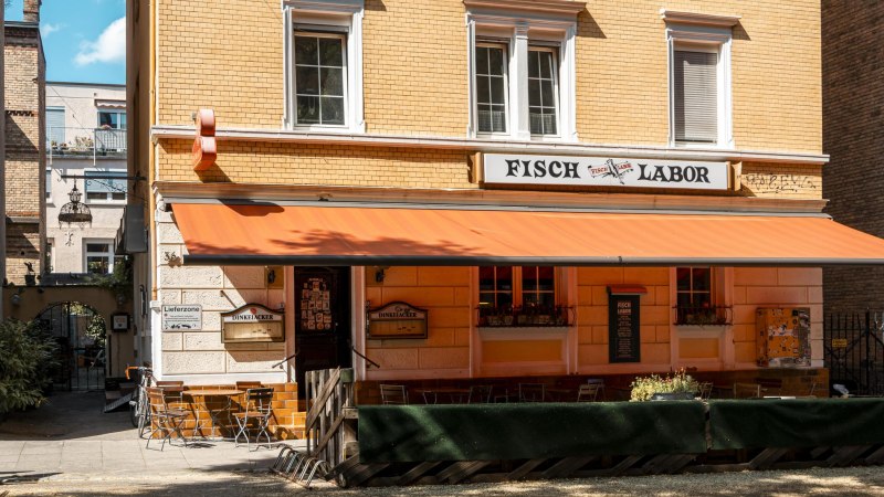 Ein Restaurant namens Fischlabor mit orangener Markise und Sitzgelegenheiten im Freien. Das Gebäude hat gelbe Ziegelwände und weiße Fensterrahmen., © SMG, Sarah Schmid Ein Restaurant namens Fischlabor mit orangener Markise und Sitzgelegenheiten im Freien. Das Gebäude hat gelbe Ziegelwände und weiße Fensterrahmen., © SMG, Sarah Schmid