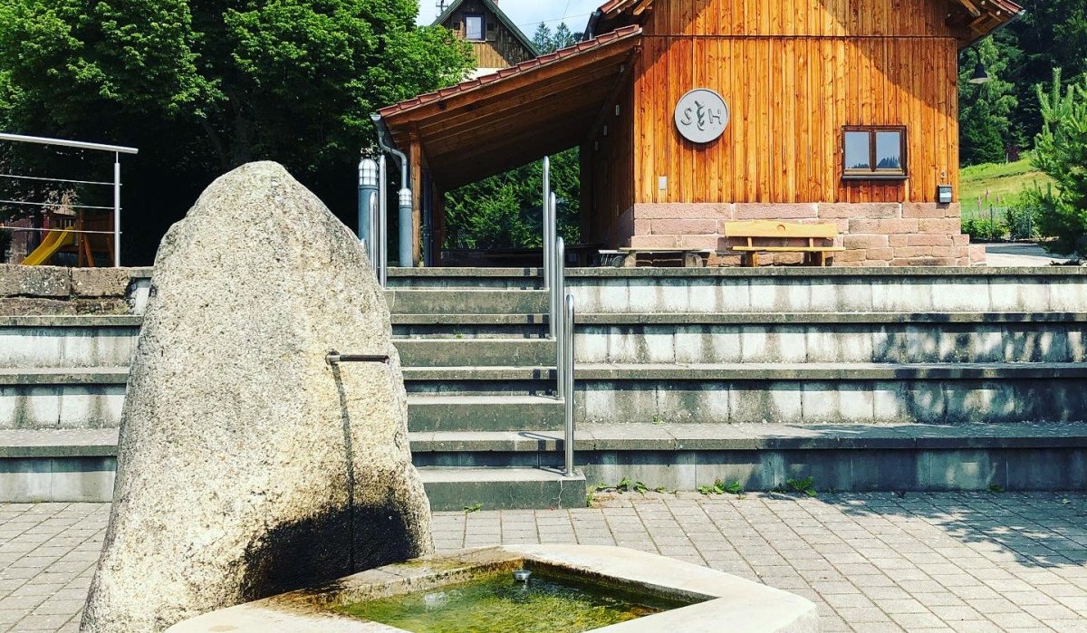 Steinbrunnen auf gepflastertem Platz, dahinter ein Holzgebäude mit Treppen und Bäumen. Klare, sonnige Atmosphäre., © Nördlicher Schwarzwald Steinbrunnen auf gepflastertem Platz, dahinter ein Holzgebäude mit Treppen und Bäumen. Klare, sonnige Atmosphäre., © Nördlicher Schwarzwald
