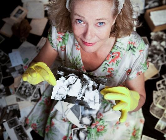 Frau in gebl&uuml;mtem Kleid und gelben Handschuhen h&auml;lt ein altes Foto, umgeben von verstreuten Fotos und Dokumenten auf dem Boden., &copy; Backnang