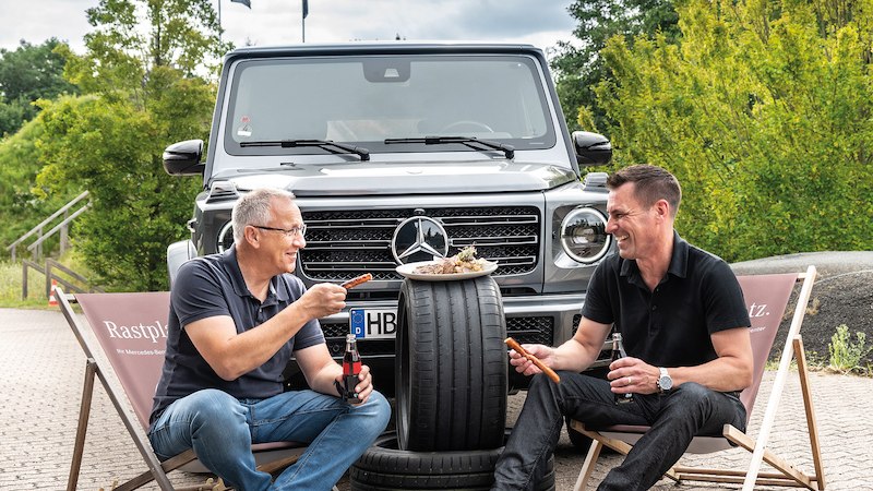 Zwei M&auml;nner sitzen auf Liegest&uuml;hlen vor einem Mercedes-Gel&auml;ndewagen und grillen auf einem Autoreifen. Sie lachen und genie&szlig;en die entspannte Atmosph&auml;re., &copy; Mercedes-Benz AG