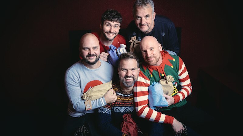 F&uuml;nf M&auml;nner in bunten Weihnachtspullovern halten Geschenkt&uuml;ten und l&auml;cheln fr&ouml;hlich in die Kamera. Die Szene wirkt festlich und herzlich., &copy; Scala Kultur Live gGmbH