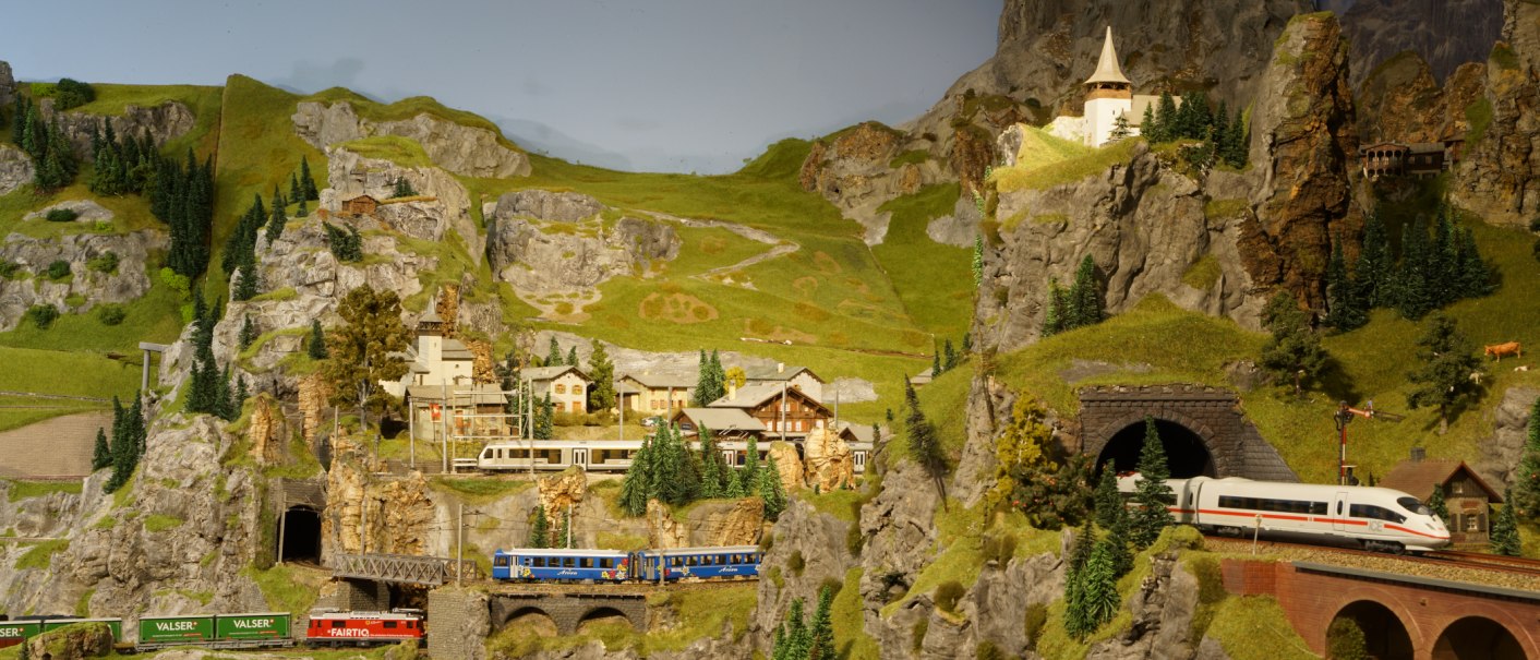 Eine detaillierte Modelleisenbahnlandschaft mit Zügen, die durch eine bergige Szenerie mit Tunneln und kleinen Gebäuden fahren., © PMW Winnenden e.V. Eine detaillierte Modelleisenbahnlandschaft mit Zügen, die durch eine bergige Szenerie mit Tunneln und kleinen Gebäuden fahren., © PMW Winnenden e.V.