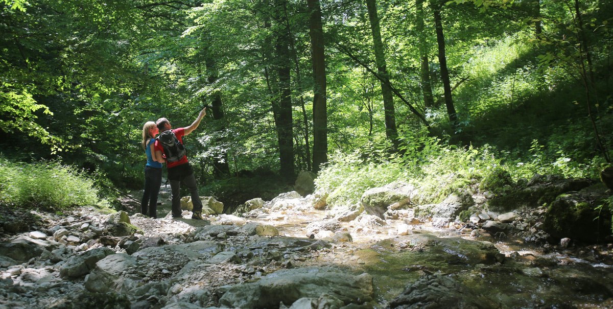 Zwei Personen stehen in einem bewaldeten Tal und machen ein Selfie. Ein kleiner Bach fließt im Vordergrund, umgeben von üppigem Grün., © Bad Urach Toruismus