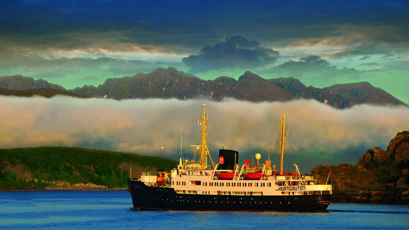 Ein Hurtigruten-Schiff fährt auf ruhigem Wasser, umgeben von Bergen und Wolken. Die Szene wirkt friedlich und malerisch., © BLICKFANG Ein Hurtigruten-Schiff fährt auf ruhigem Wasser, umgeben von Bergen und Wolken. Die Szene wirkt friedlich und malerisch., © BLICKFANG
