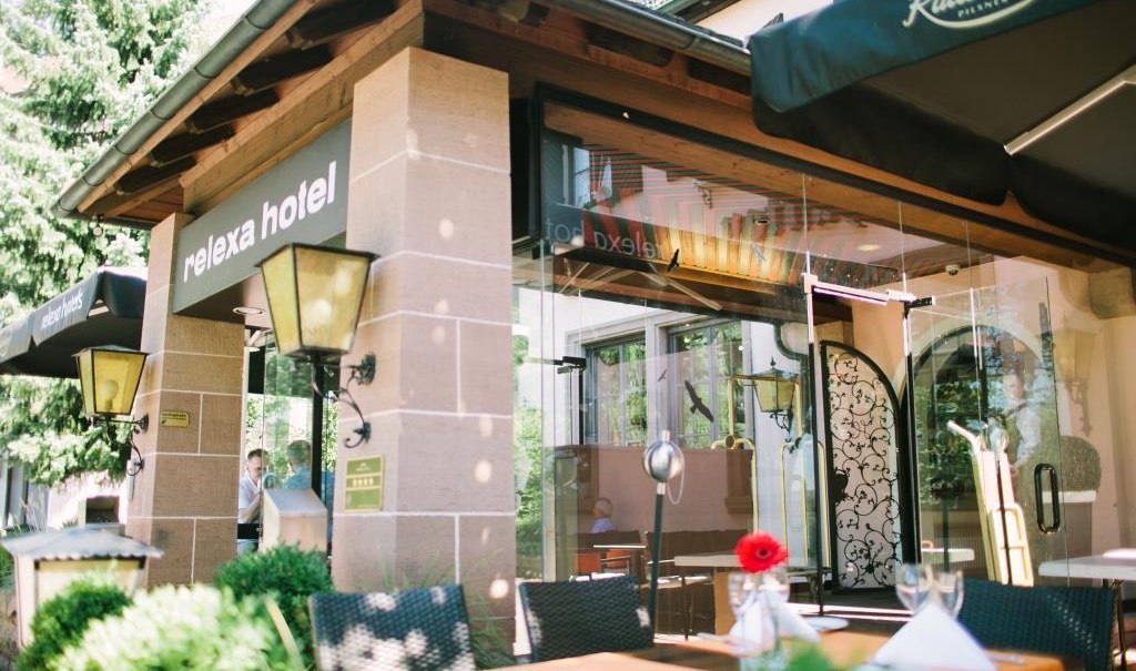 Eingang des relexa hotels mit Glasfront und Terrasse. Tische mit weißen Tischdecken und Blumen, umgeben von grünen Pflanzen., © relexa Waldhotel Schatten Eingang des relexa hotels mit Glasfront und Terrasse. Tische mit weißen Tischdecken und Blumen, umgeben von grünen Pflanzen., © relexa Waldhotel Schatten