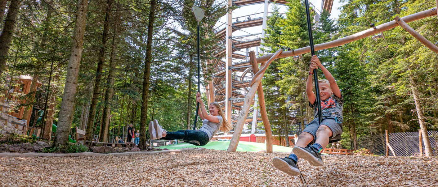 Zwei Kinder schaukeln fröhlich im Abenteuerwald Sommerberg. Im Hintergrund ist ein großer Holzpfad zu sehen, umgeben von hohen Bäumen., © Erlebnis Akademie AG Zwei Kinder schaukeln fröhlich im Abenteuerwald Sommerberg. Im Hintergrund ist ein großer Holzpfad zu sehen, umgeben von hohen Bäumen., © Erlebnis Akademie AG
