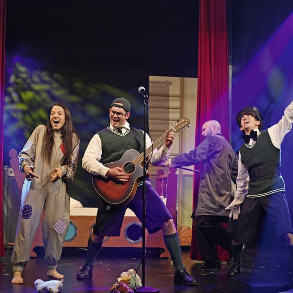 Vier Darsteller auf einer Bühne, einer spielt Gitarre, die anderen tanzen und lachen. Bunte Lichter und Requisiten im Hintergrund., © Stadtverwaltung Schorndorf