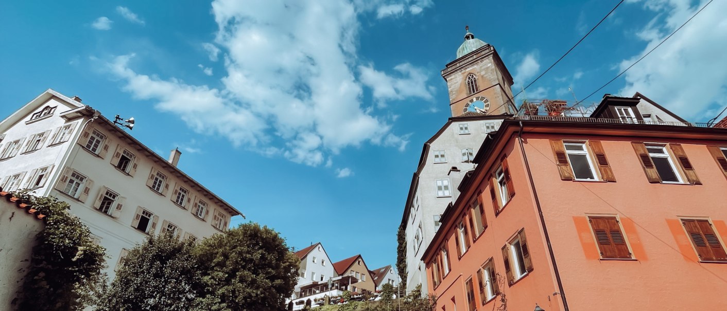 Ein Café mit roten Wänden und Außensitzbereich auf Kopfsteinpflaster. Im Hintergrund ein Kirchturm und blauer Himmel mit Wolken., © Jiggis