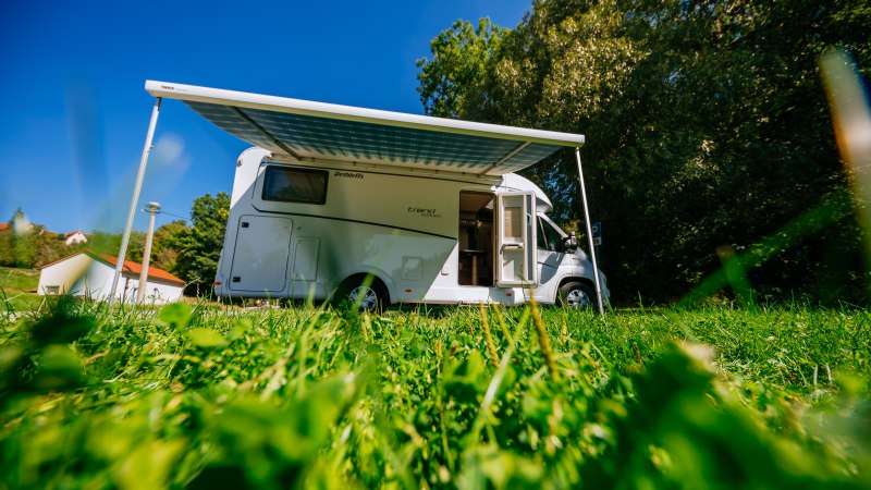 Ein Wohnmobil mit ausgefahrener Markise steht auf einer gr&uuml;nen Wiese unter blauem Himmel. Im Hintergrund sind B&auml;ume und ein Geb&auml;ude zu sehen., &copy; SMG, Thomas Niederm&uuml;ller