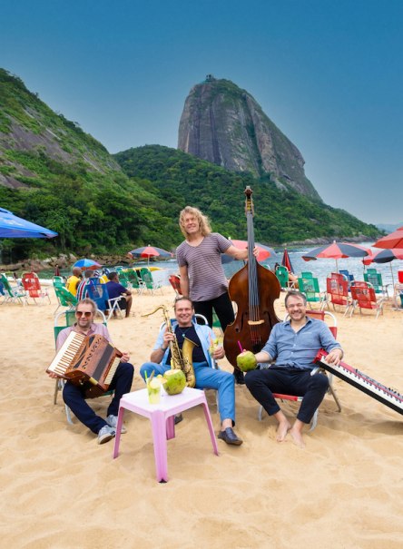 Vier Musiker sitzen am Strand mit Instrumenten und Getränken. Im Hintergrund ist ein großer Felsen zu sehen. Der Himmel ist klar und blau., © Stadt Leinfelden-Echterdingen, Kulturamt