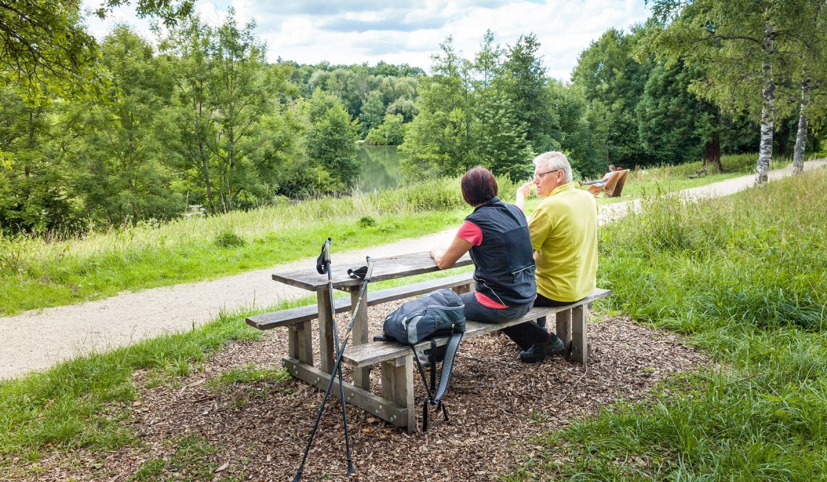 Zwei Personen sitzen an einem Holztisch im Grünen, mit Wanderstöcken und Rucksack. Sie blicken auf einen See, umgeben von Bäumen und einem Weg., © hochgehberge Zwei Personen sitzen an einem Holztisch im Grünen, mit Wanderstöcken und Rucksack. Sie blicken auf einen See, umgeben von Bäumen und einem Weg., © hochgehberge