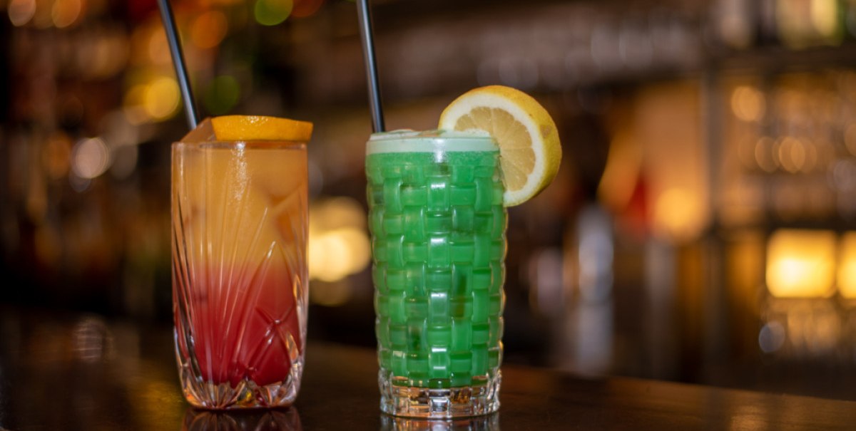 Zwei bunte Cocktails auf einer Bar, einer orange-rot, der andere grün, mit Zitronenscheiben und Strohhalmen., © Café Bar Zill