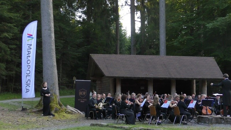 Ein Orchester spielt ein Konzert im Freien vor einer Holzh&uuml;tte im Wald. Eine Fahne mit der Aufschrift 'Małopolska' steht links., &copy; Touristik Bad Wildbad GmbH