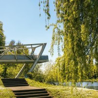 Eine moderne Brücke erstreckt sich über einen Fluss, umgeben von üppigem Grün und Bäumen unter einem klaren blauen Himmel., © Stuttgart-Marketing GmbH, Sarah Schmid Eine moderne Brücke erstreckt sich über einen Fluss, umgeben von üppigem Grün und Bäumen unter einem klaren blauen Himmel., © Stuttgart-Marketing GmbH, Sarah Schmid