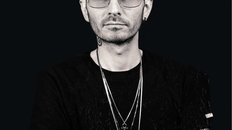 Person mit Glatze, Sonnenbrille und tätowierten Armen vor schwarzem Hintergrund. Sie trägt ein schwarzes T-Shirt und mehrere Halsketten., © Expedition Erde Person mit Glatze, Sonnenbrille und tätowierten Armen vor schwarzem Hintergrund. Sie trägt ein schwarzes T-Shirt und mehrere Halsketten., © Expedition Erde