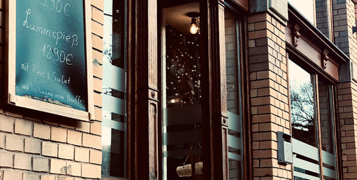 Eingang eines Restaurants mit Backsteinfassade. Eine Tafel zeigt Preise für Hähnchen- und Lammspieße. Vor der Tür stehen Tische mit Blumenkübeln., © Bady's