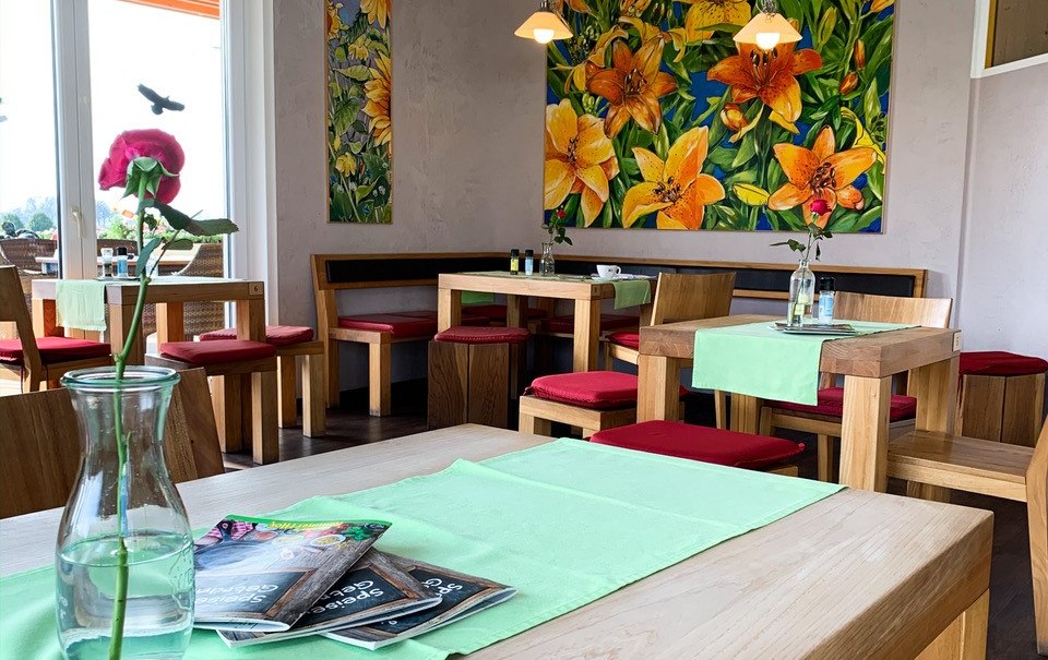 Gemütlicher Café-Innenraum mit Holztischen, roten Sitzpolstern, grünen Tischläufern und farbenfrohen Blumenbildern an der Wand., © Engel/ Rammerthof