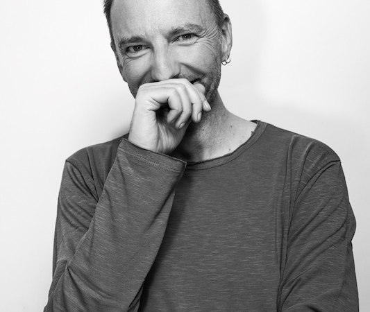 Ein Mann in einem langärmeligen Shirt lächelt und hält seine Hand vor den Mund. Schwarz-weiß Fotografie., © Kulturhaus Schwanen