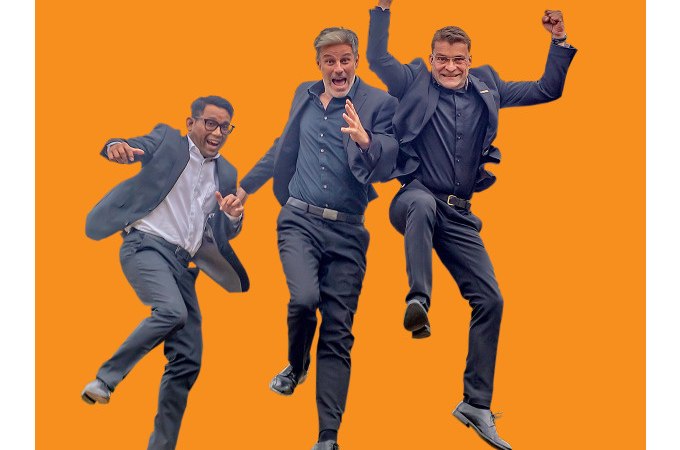 Drei M&auml;nner in Anz&uuml;gen springen fr&ouml;hlich in die Luft vor einem orangefarbenen Hintergrund. Sie wirken energiegeladen und begeistert., &copy; Stadt Murrhardt