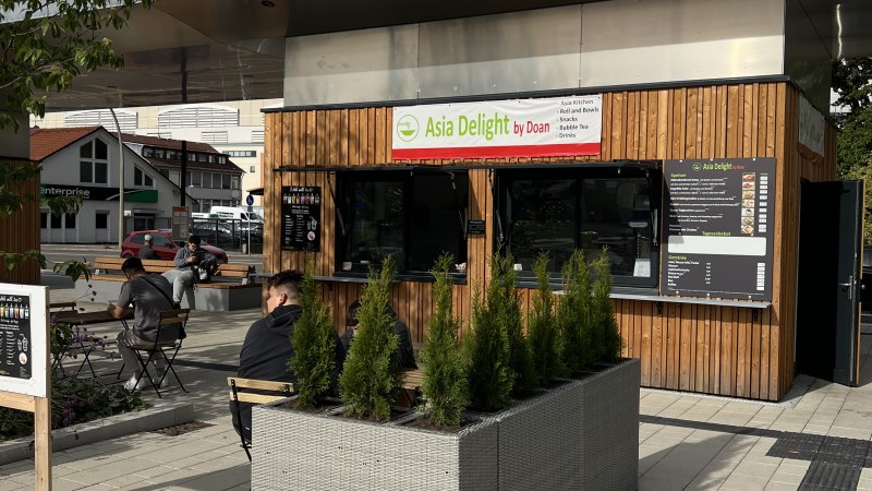 Kleiner Kiosk 'Asia Delight by Doan' mit Holzverkleidung und Sitzbereich im Freien. Menschen sitzen an Tischen, umgeben von Pflanzen., &copy; Asia Delight GmbH