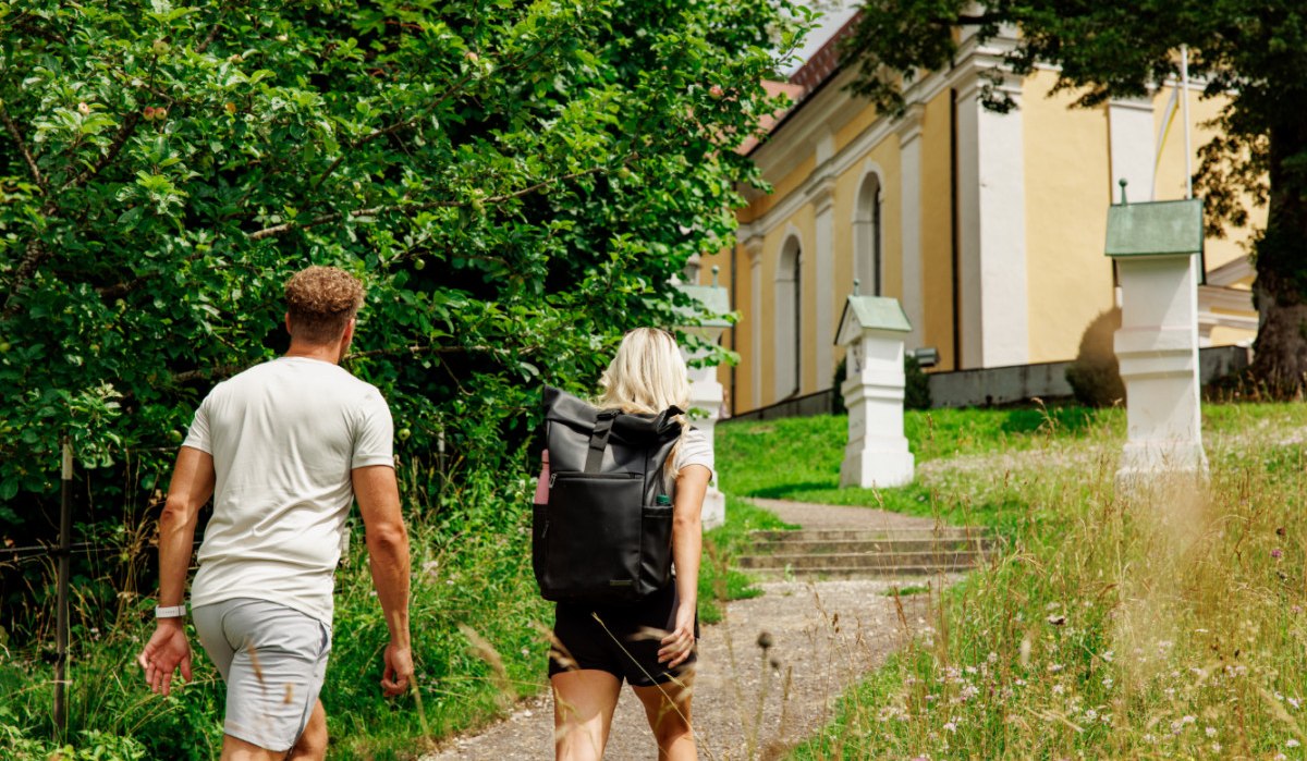 Zwei Personen wandern auf einem Weg neben einer gelben Kirche, umgeben von grünen Bäumen und Wiesen., © Landkreis Göppingen