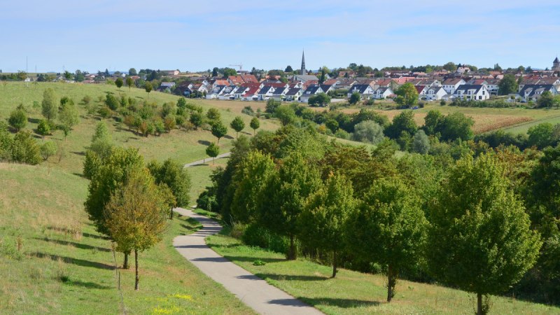 Ein gewundener Weg führt durch eine grüne Landschaft mit Bäumen. Im Hintergrund ist eine Siedlung mit Häusern und einer Kirche sichtbar., © Remstal Tourismus e.V.